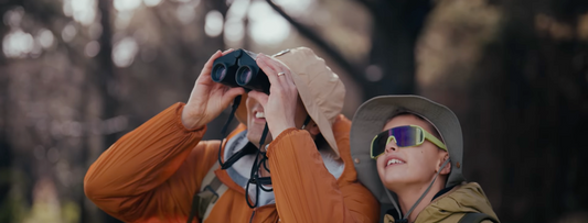 A man in an orange jacket uses Solvia 8x32 binoculars next to a child wearing a hat and reflective sunglasses; both are observing something together outdoors.