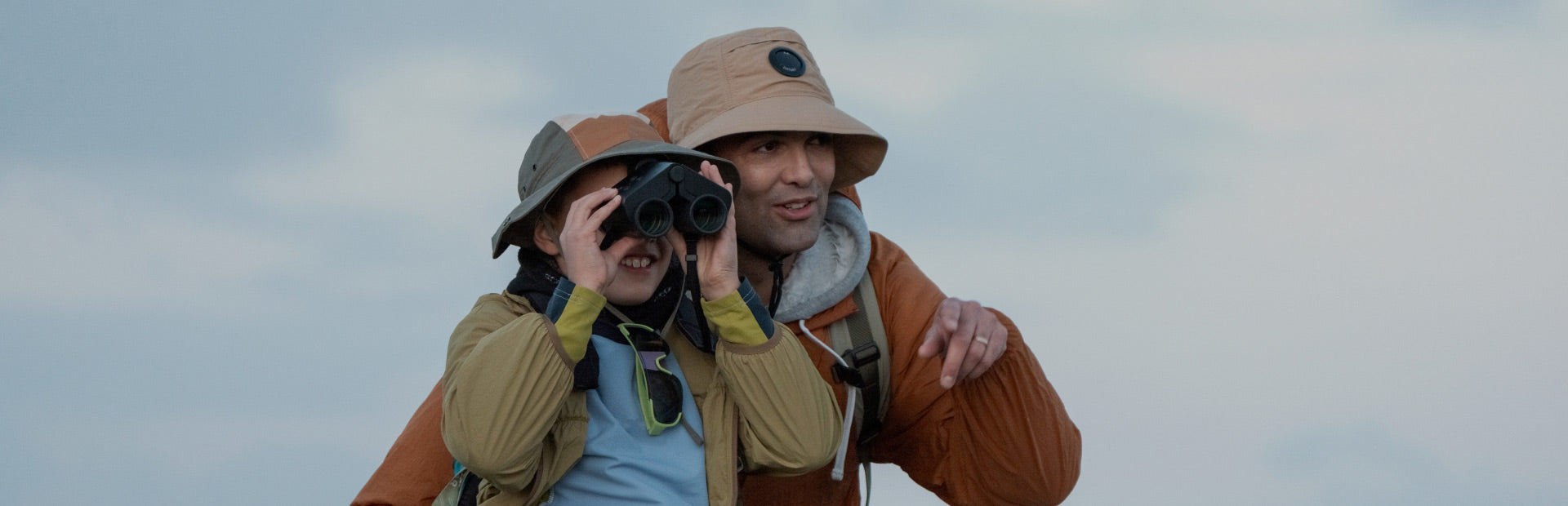 Two people in outdoor gear with Solvia ED 8x32 compact digital binoculars against a cloudy sky.