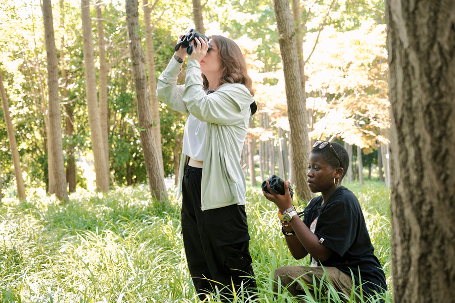 Two birdwatchers using binoculars in a wooded park during the Matataxplore Migration Challenge. One standing and looking through binoculars while the other crouches holding a camera. Tall trees and grass are visible in the background.