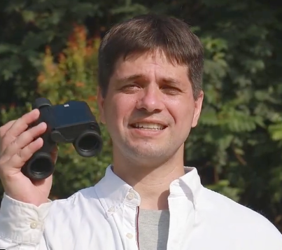 A man outdoors holding Solvia 8x32 AI binoculars, wearing a white button-up shirt over a gray undershirt, with green foliage in the background.